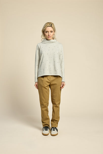 A woman wearing the Reminder Sweater by Cokluch in Natural, with a high twisted neck, darts and the shoulders, long sleeves, and a loose fit. She is wearing it with yellow pants and standing in front of a beige background. 