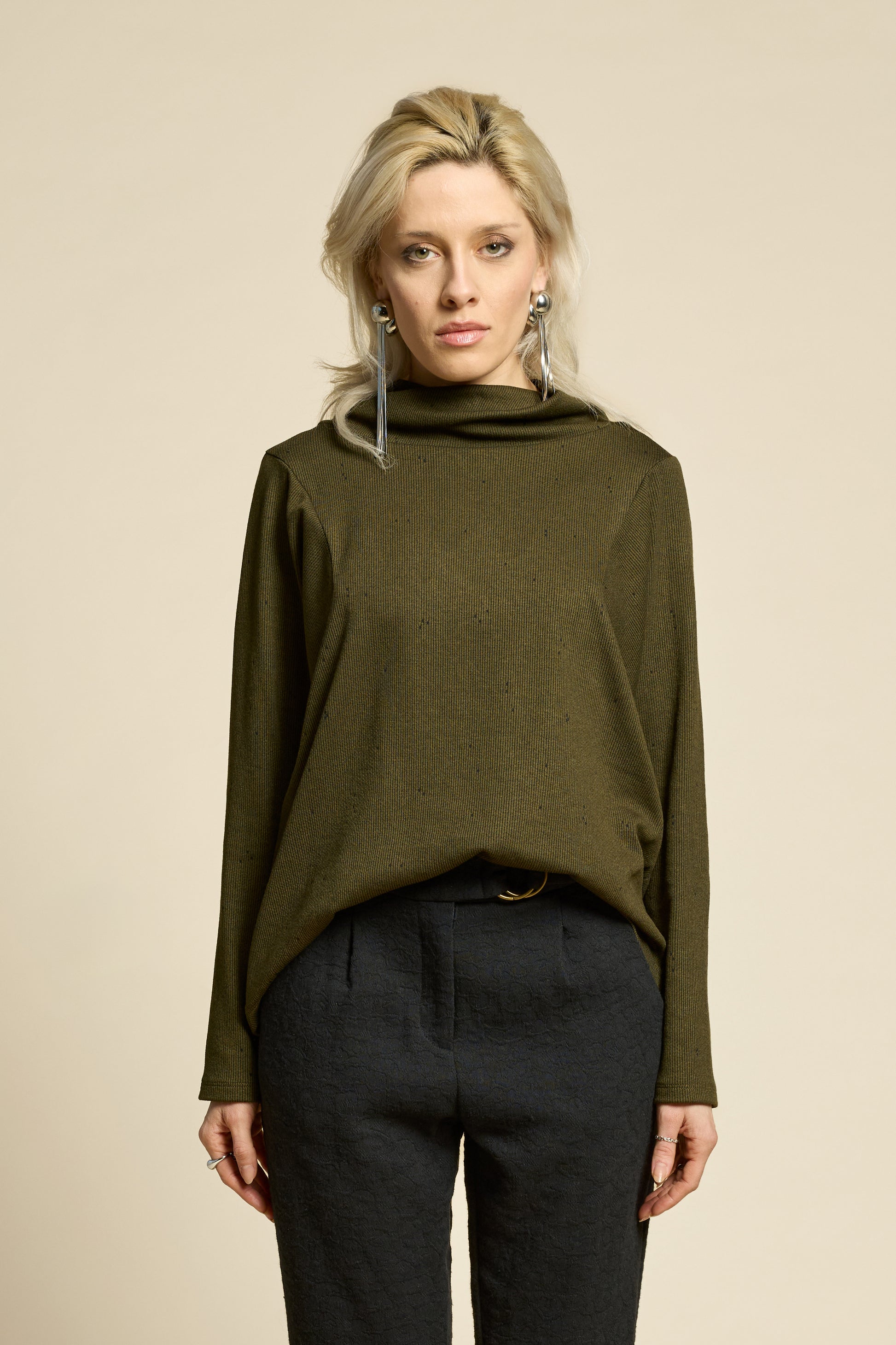 Waist up view of a woman wearing the Reminder Sweater by Cokluch in Olive, with a high twisted neck, darts and the shoulders, long sleeves, and a loose fit. She is wearing it with black pants and standing in front of a beige background. 