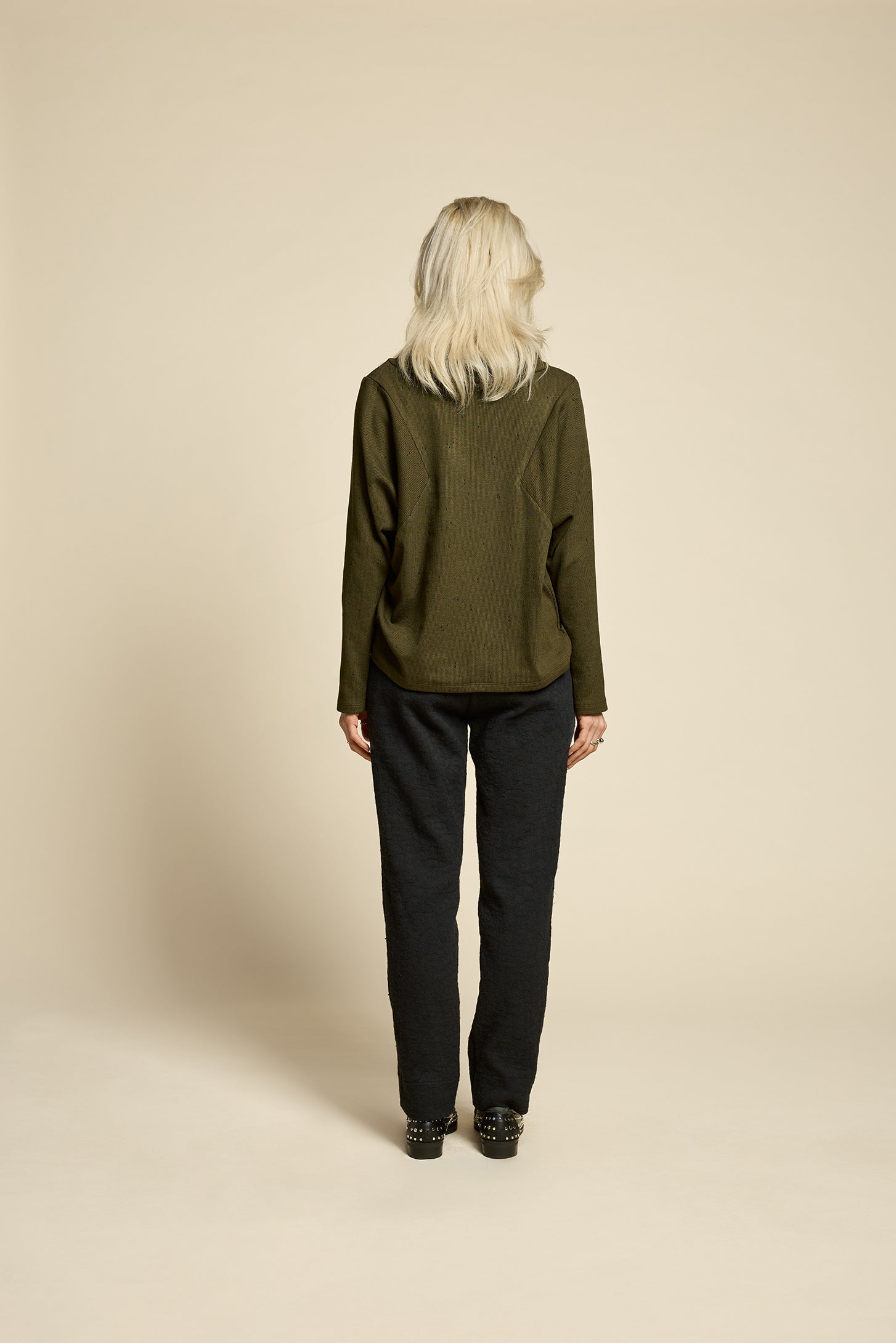 Back view of a woman wearing the Reminder Sweater by Cokluch in Olive, with a high twisted neck, darts and the shoulders, long sleeves, and a loose fit. She is wearing it with black pants and standing in front of a beige background. 