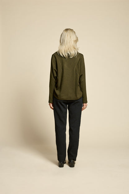 Back view of a woman wearing the Reminder Sweater by Cokluch in Olive, with a high twisted neck, darts and the shoulders, long sleeves, and a loose fit. She is wearing it with black pants and standing in front of a beige background. 