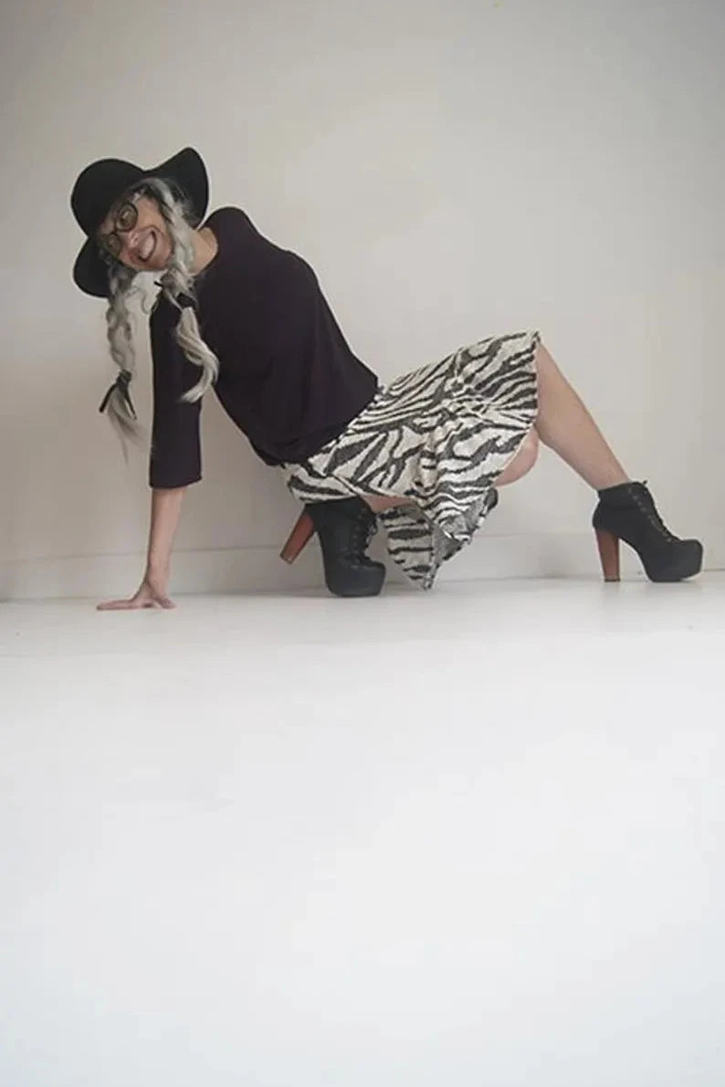 A woman wearing the knee-length Marvel Skirt by SI Design in Ivory/Black, featuring a pull-on waist, a swirling pattern, and a gathered panel at the bottom. She is wearing it with a black top and is kneeling in front of a white background. 