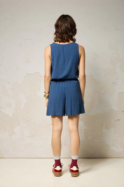Back view of a woman wearing the Atman Shorts by Cokluch in Denim blue, mid-thigh length shorts with a high elastic waist and an attached ring belt. She is wearing them with a matching top and standing in front of a plain background. 