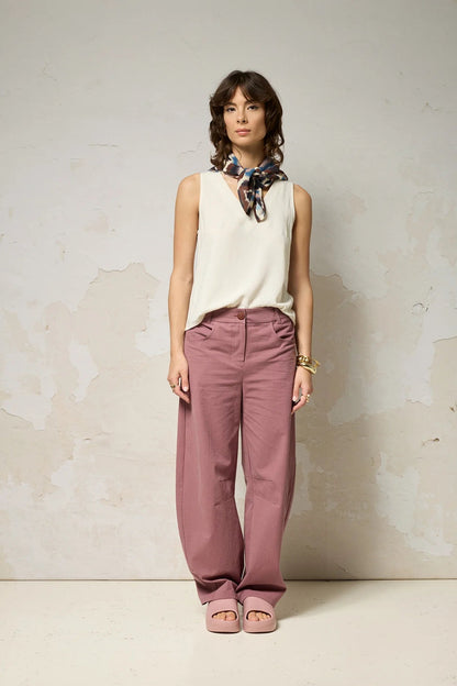 A woman wearing the Sita Top by Cokluch in Natural a sleeveless V-neck tank with a loose fit and wide straps. She is wearing it with pink pants and a patterned scarf against a plain background