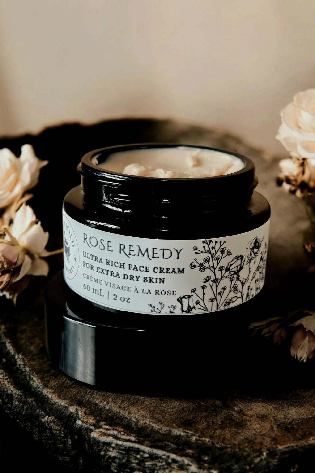 A jar of 'Rose Remedy' ultra rich face cream by Stay Wild Apothecary is shown with flowers on a stone surface