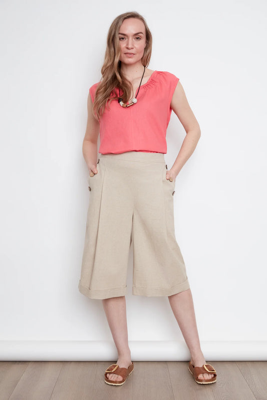 A woman wearing the Sabrina Top by Julei in coral, with raglan cap sleeves, a notched v-neck with pleat details, and a pleated back yoke. She is wearing it with the Isadora Capri pants in Natural and standing against a white background