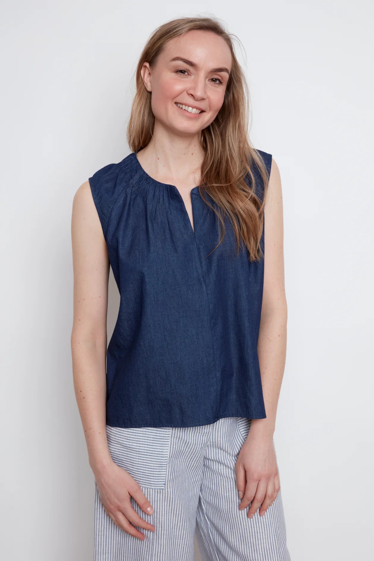 A woman wearing the Sabrina Top by Julei in Denim, with raglan cap sleeves, a notched v-neck with pleat details, and a pleated back yoke. She is wearing it with striped pants and standing against a white background
