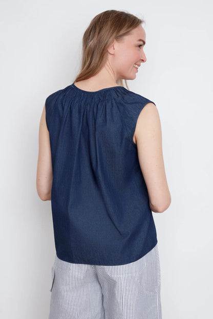 Back view of a woman wearing the Sabrina Top by Julei in Denim, with raglan cap sleeves, a notched v-neck with pleat details, and a pleated back yoke. She is wearing it with striped pants and standing against a white background