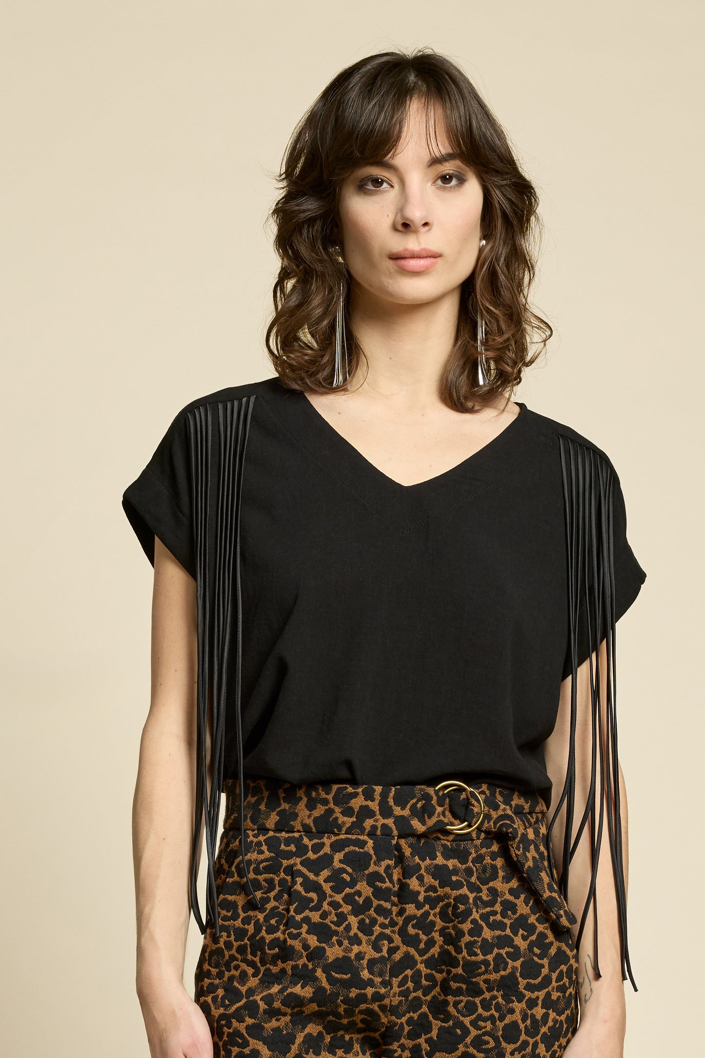 A woman wearing the Shout Top by Cokluch in Harper Black, with fringe at the shoulders, short extended sleeves, and a V-neck. She is wearing it with the Breeze Pants in Cappucino Leopard print and standing against on a beige background. 