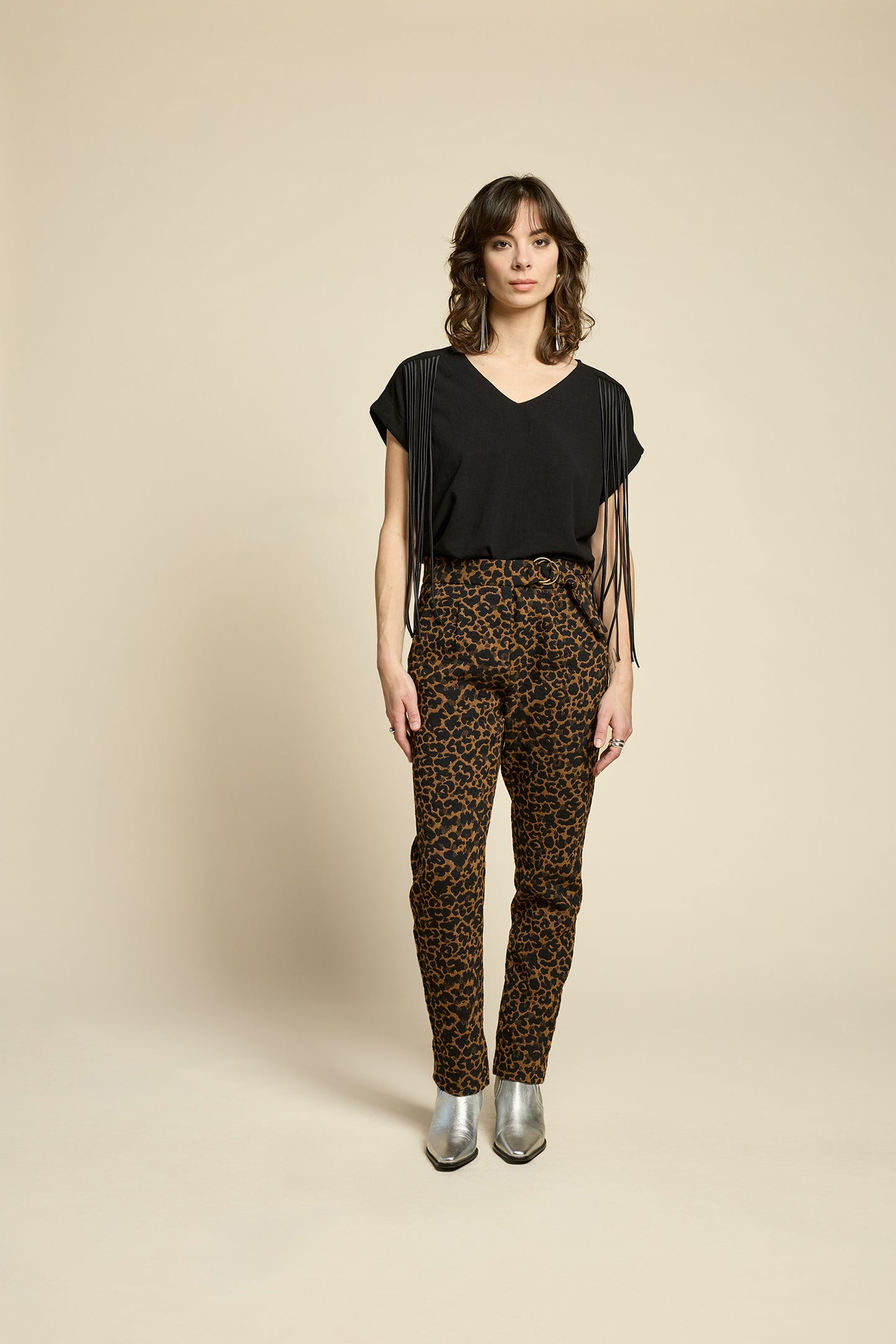 A woman wearing the Shout Top by Cokluch in Harper Black, with fringe at the shoulders, short extended sleeves, and a V-neck. She is wearing it with the Breeze Pants in Cappucino Leopard print and standing against on a beige background. 