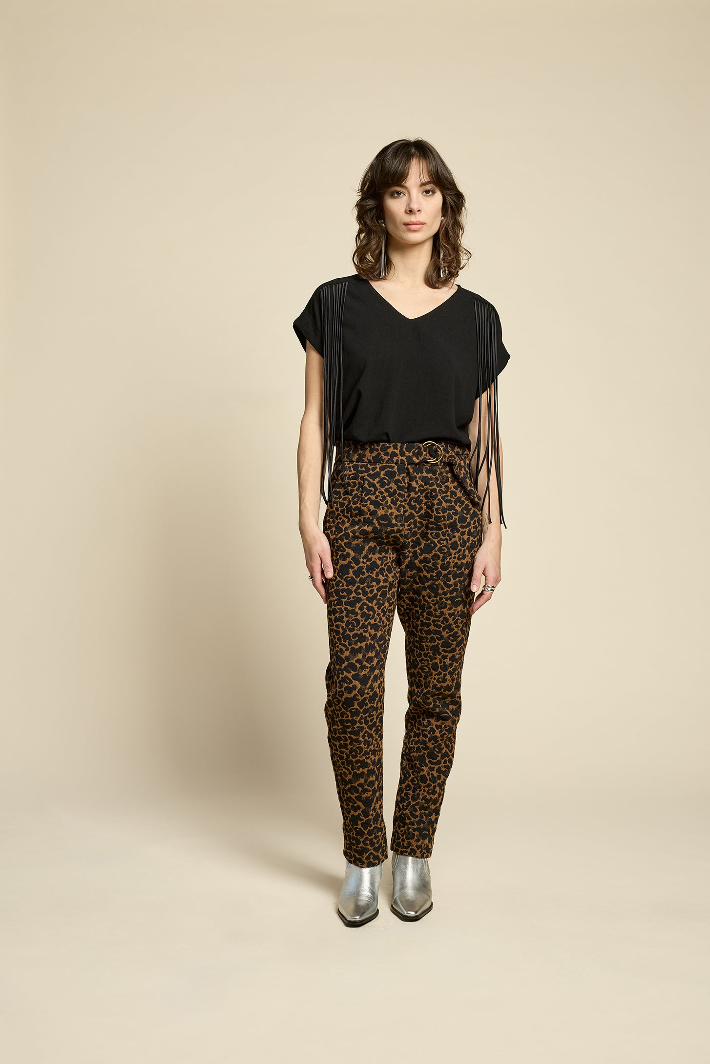 A woman wearing the Shout Top by Cokluch in Harper Black, with fringe at the shoulders, short extended sleeves, and a V-neck. She is wearing it with the Breeze Pants in Cappucino Leopard print and standing against on a beige background. 