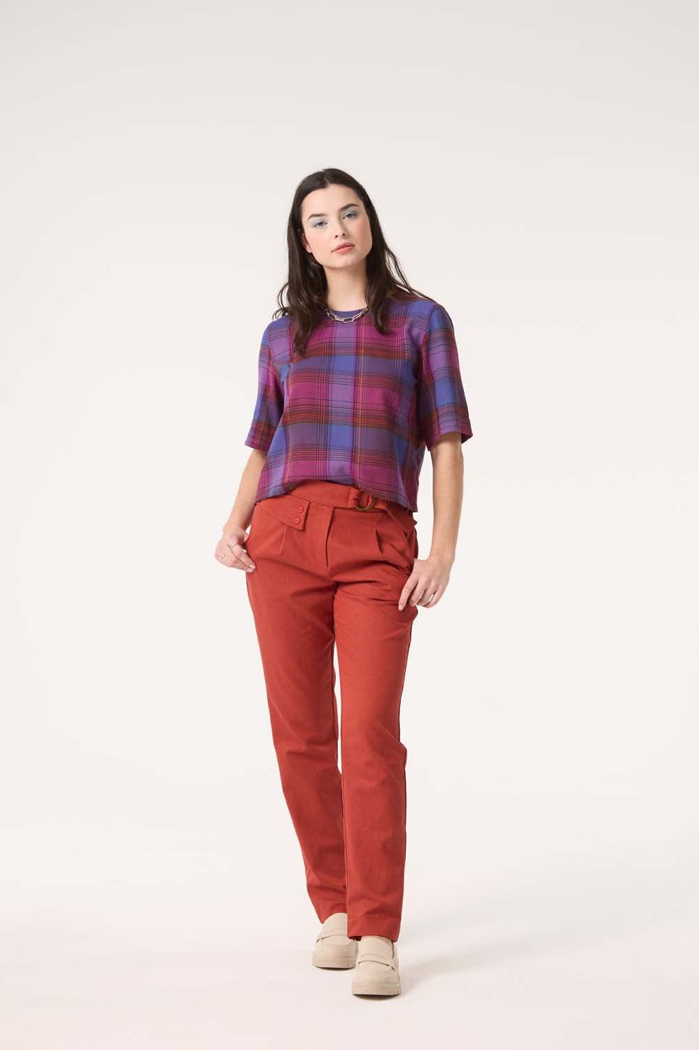 A woman wearing the Sideral Top by Cokluch in Purple Plaid with orange pants, standing in front of white background 