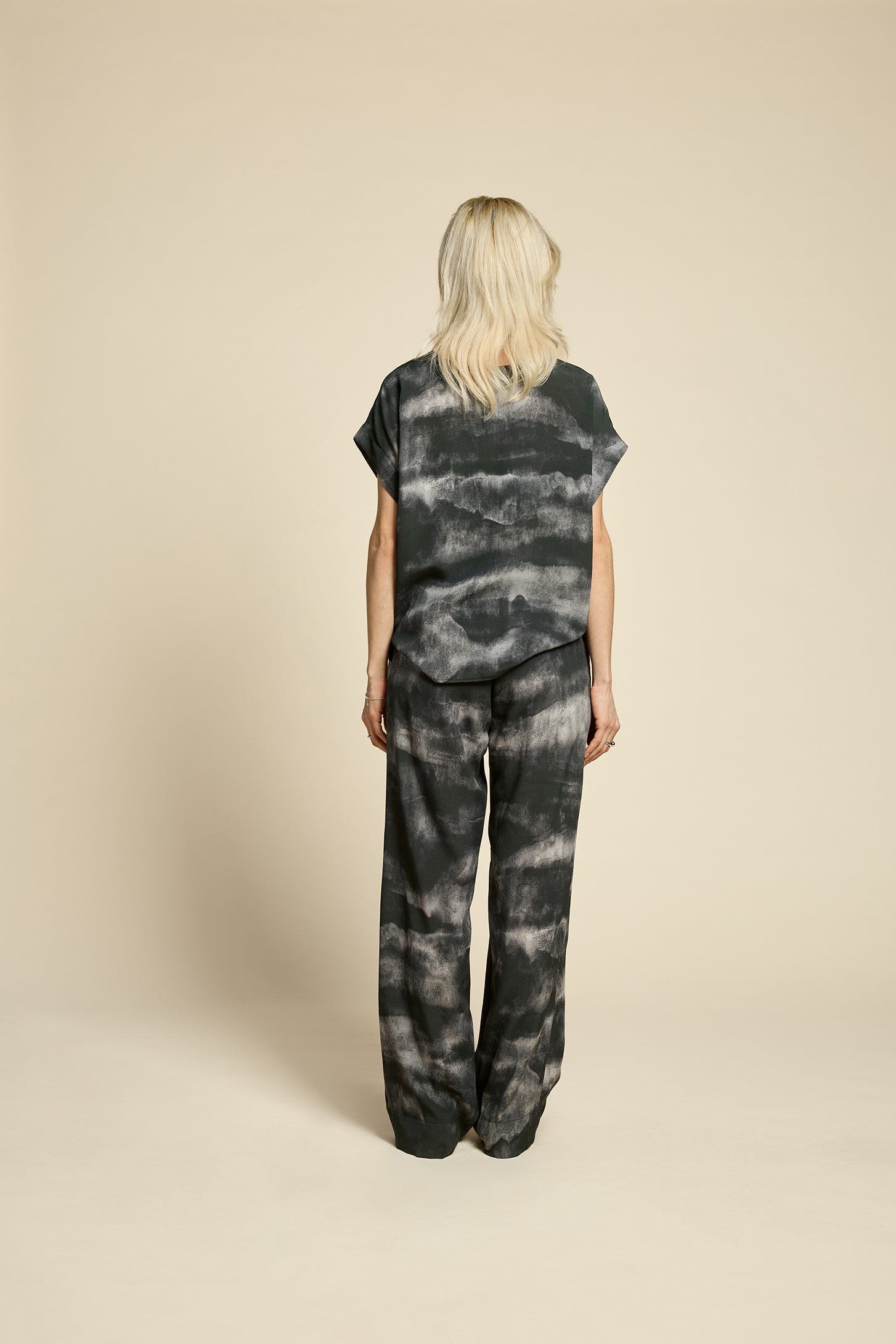 Back view of a woman wearing the Silence Top by Cokluch in Mist Black, with a round neck, short Magyar sleeves, and a loose fit. She is wearing it with matching pants and standing in front of a beige background. 