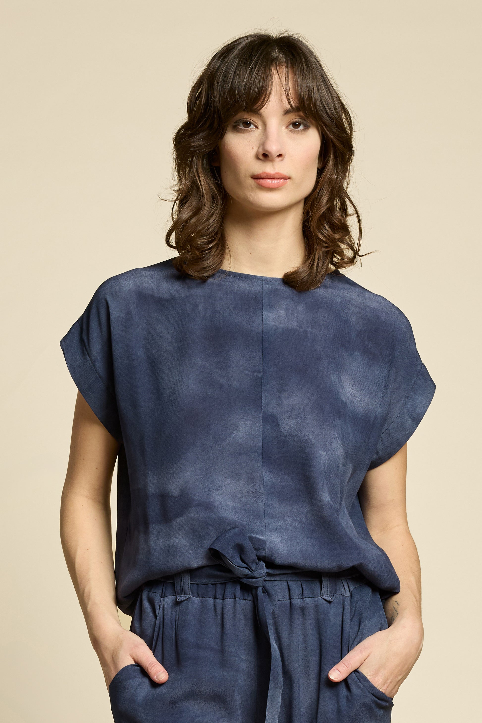 Waist up view of a woman wearing the Silence Top by Cokluch in Mist Twilight, with a round neck, short Magyar sleeves, and a loose fit. She is wearing it with matching pants and standing in front of a beige background. 