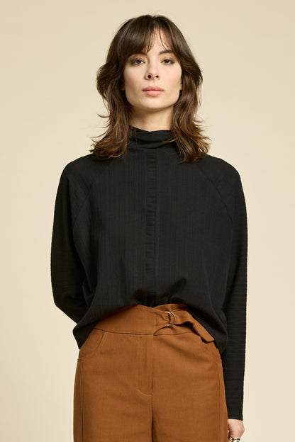 Waist up view of a woman wearing the Souvenir Top by Cokluch in Black, with a twisted turtleneck, ribbed fabric with a vertical band on the front. She's wearing it with yellow pants and standing in front of a beige background. 