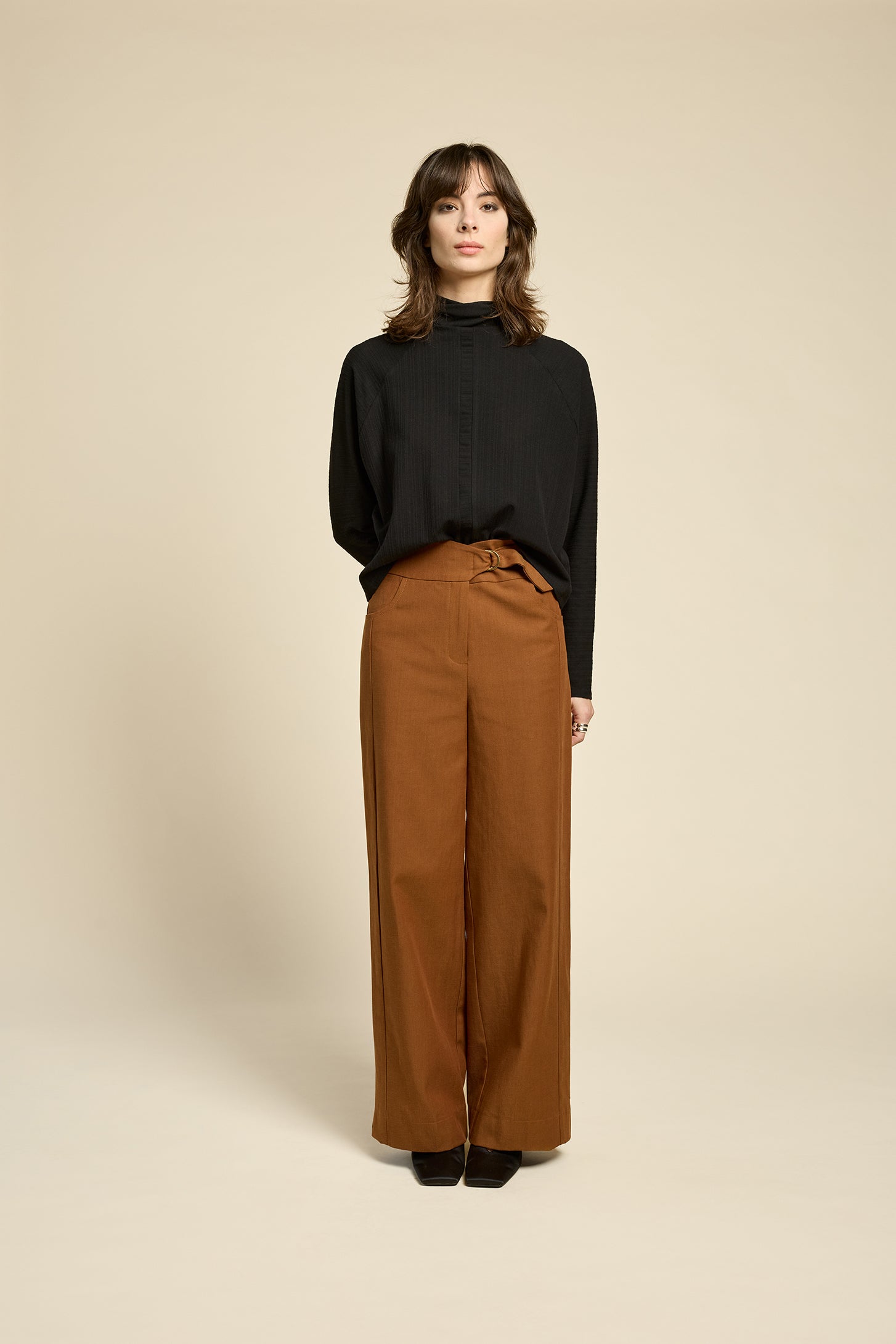 A woman wearing the Souvenir Top by Cokluch in Black, with a twisted turtleneck, ribbed fabric with a vertical band on the front. She's wearing it with brown pants and standing in front of a beige background. 