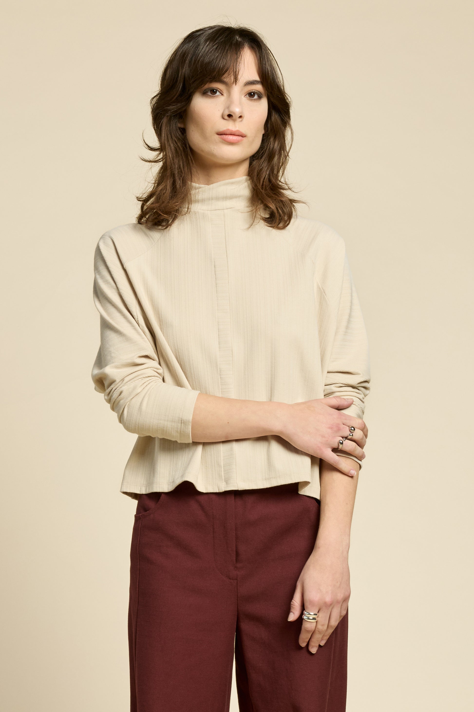 Waist up view of a woman wearing the Souvenir Top by Cokluch in Sand, with a twisted turtleneck, ribbed fabric with a vertical band on the front. She's wearing it with burgundy pants and standing in front of a beige background. 
