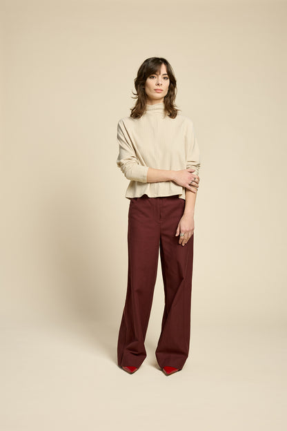 A woman wearing the Souvenir Top by Cokluch in Sand, with a twisted turtleneck, ribbed fabric with a vertical band on the front. She's wearing it with burgundy pants and standing in front of a beige background. 