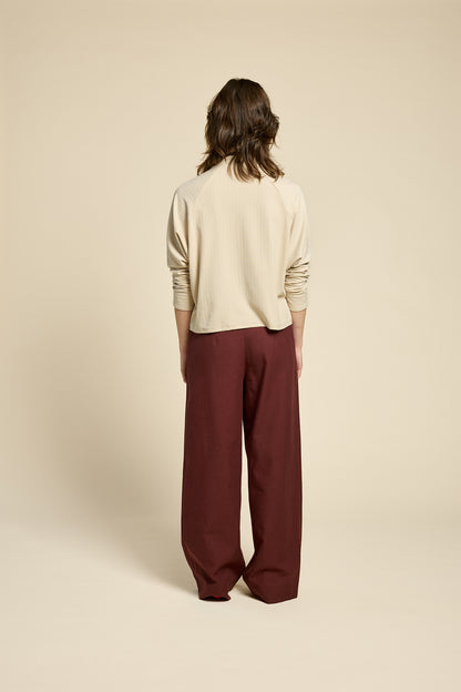 Back view of a woman wearing the Souvenir Top by Cokluch in Sand, with a twisted turtleneck, ribbed fabric with a vertical band on the front. She's wearing it with burgundy pants and standing in front of a beige background. 