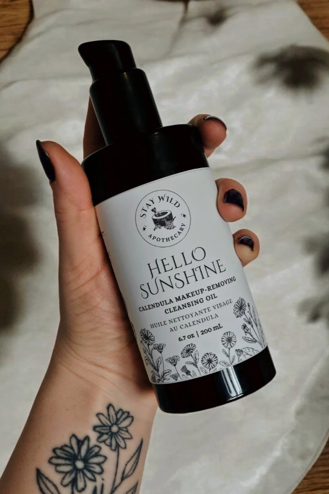 Hand holding a bottle of 'Hello Sunshine' cleansing oil by Stay Wild Apothecary with a blurred background