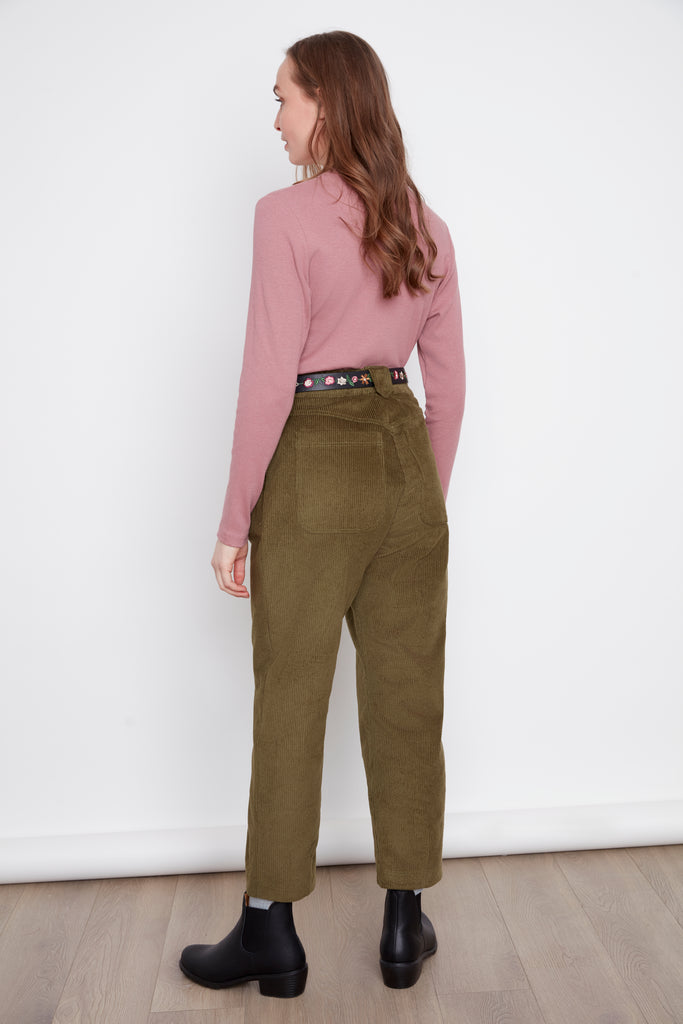 Back view of a woman wearing the Melody Mockneck Top by Julie in Rosewood, a ribbed knit top with a short cross-over collar and long sleeves. She is wearing it with green corduroy pants  against a white background.
