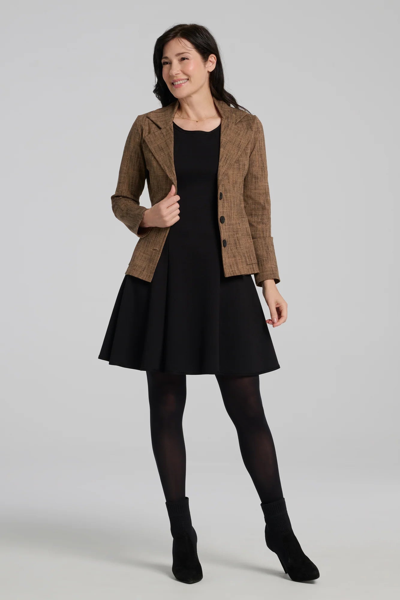 A woman wearing the Toscana Dress by Kollontai in Black, a sleeveless piece with a round neck, princess seams, and fit and flare shape with a full skirt that hits above the knee. She is wearing it with a brown blazer and standing against a grey background.