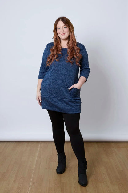 A woman wearing the Brooke Tunic by Tangente in Indigo, with 3/4 sleeves and a fitted band at the bottom. She is wearing it with black leggings and standing against a white wall. 