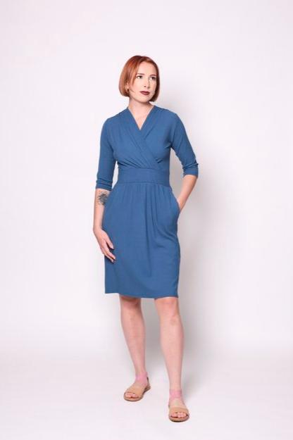 A woman wearing the Camille Dress by Tangente in Blue, featuring a cross-over neckline, narrow waistband, knee length skirt, 3/4 sleeves, and side pockets, in ribbed bamboo knit. She is standing in front of white background 