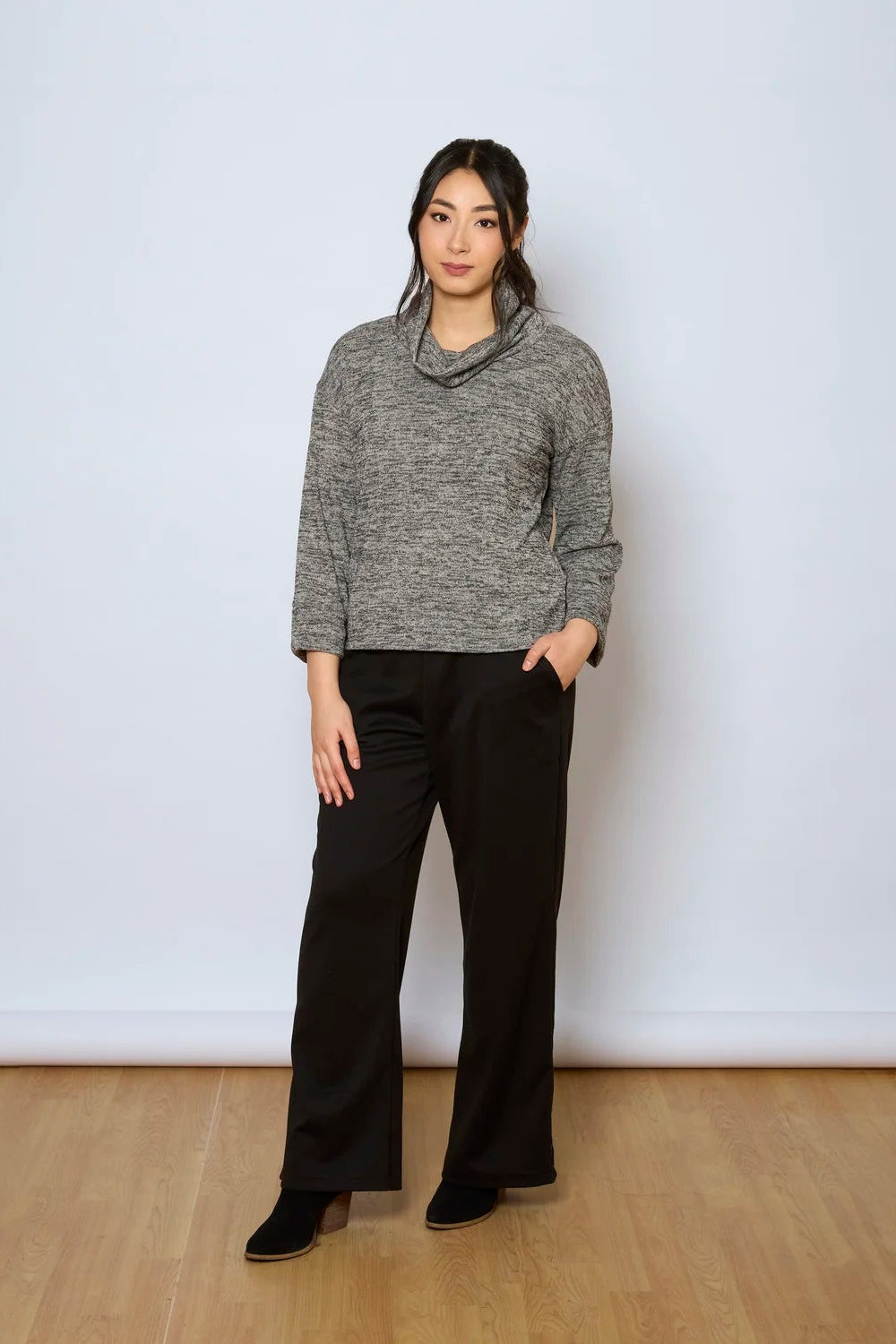 A woman wearing the Jill Pants by Tangente in Black, wide-legged ankle length pants with large pockets. She is wearing them with a grey sweater against a white background.