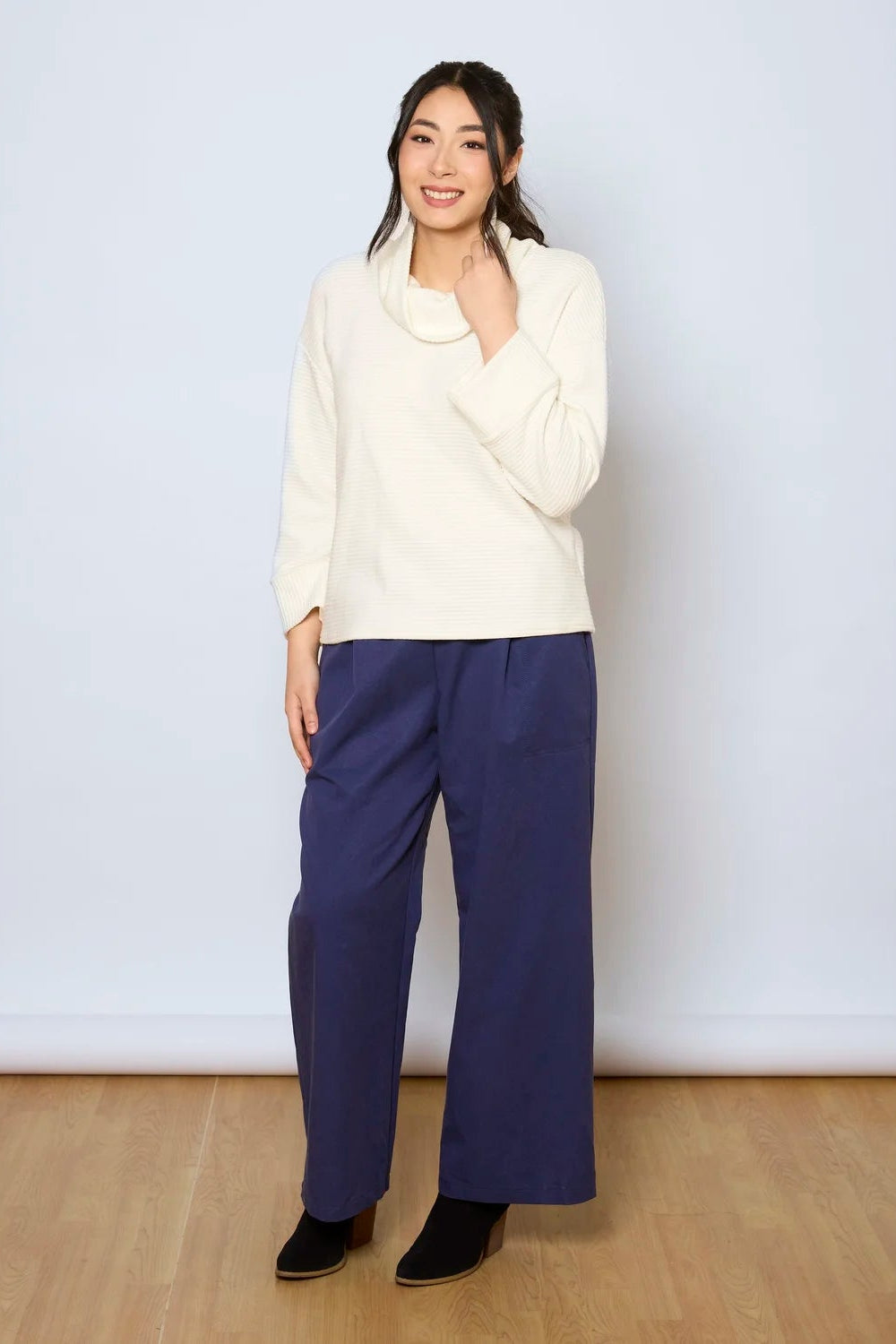 A woman wearing the Jill Pants by Tangente in Denim, wide-legged ankle length pants with large pockets. She is wearing them with a white sweater against a white background.