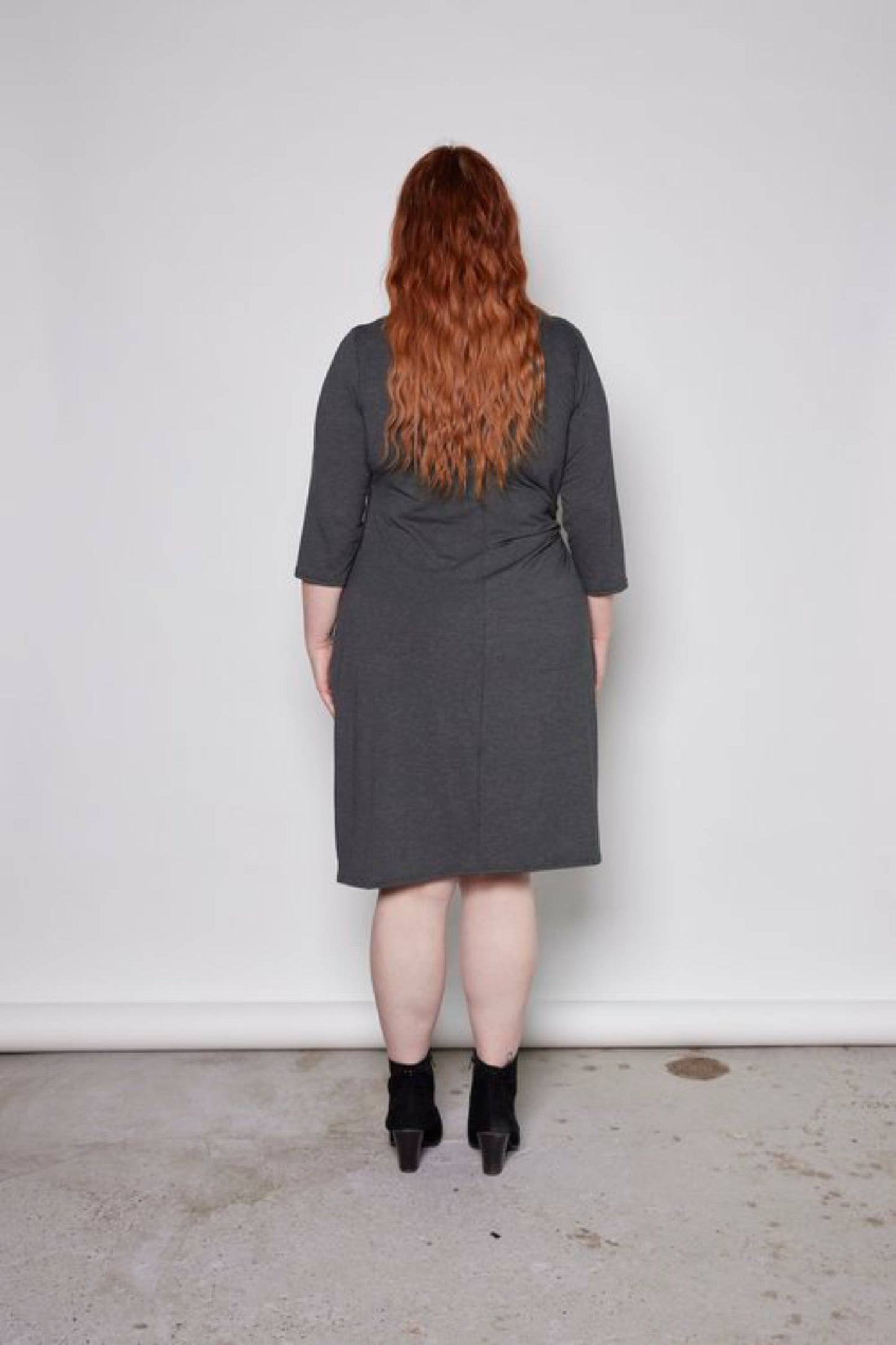 Back view of a woman wearing the Mariela Dress by Tangente in Charcoal, standing in front of a white background 