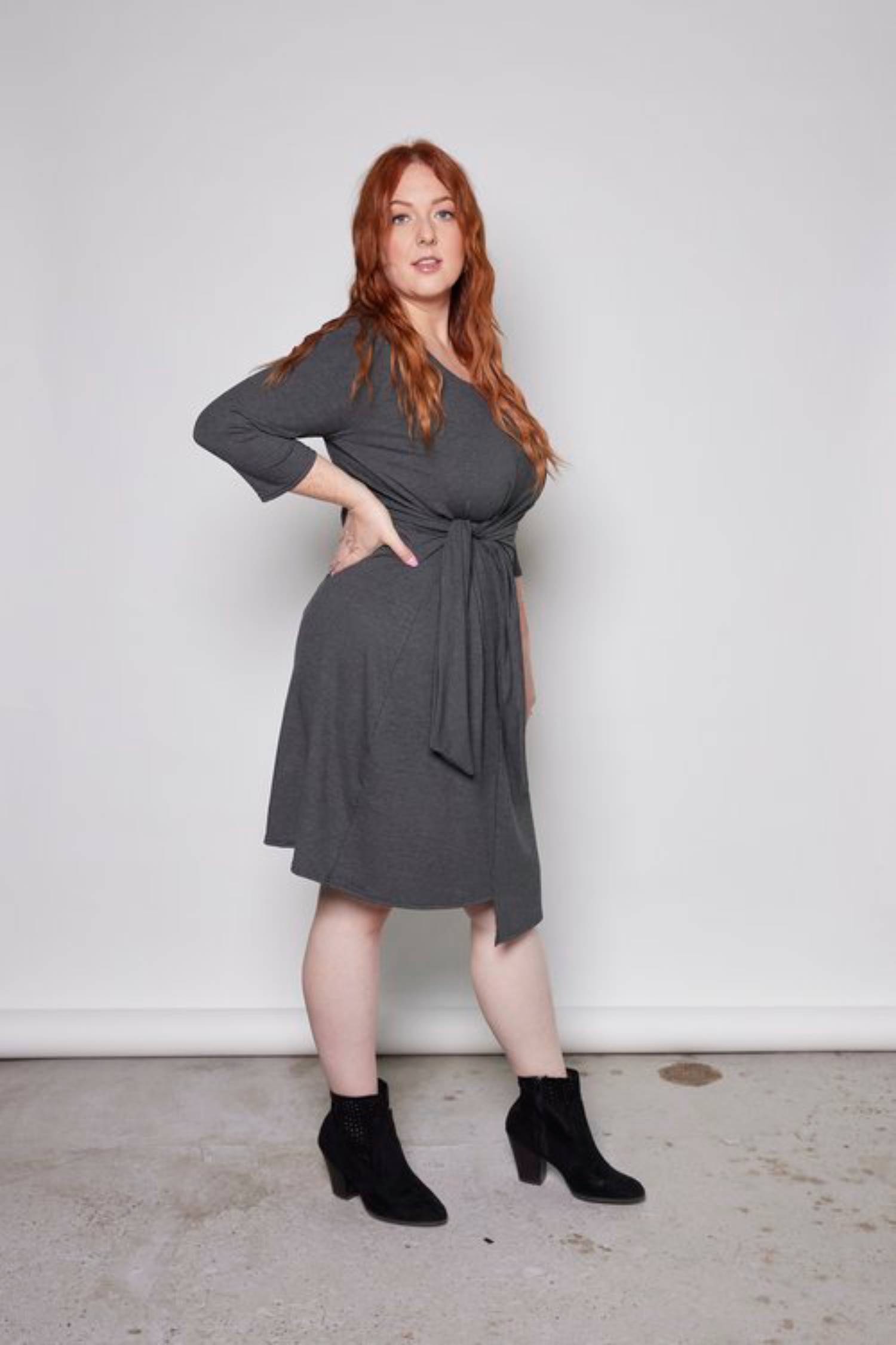 Side view of a woman wearing the Mariela Dress by Tangente in Charcoal, standing in front of a white background 