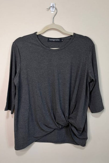 The Tangente Reese Top in Charcoal, with elbow length sleeves, a twist  at the front hem and a round neck, is shown hanging in front of a beige wall 