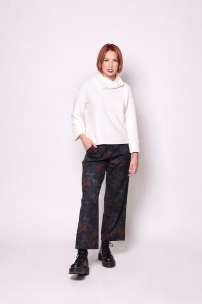 A woman wearing the Sidney Top by Tangente in Off-white, a thick ribbed knit sweater with a cowl neck, long cuffed sleeves, and a slightly boxy fit. She's wearing it with floral pants and standing in front of a white background. 
