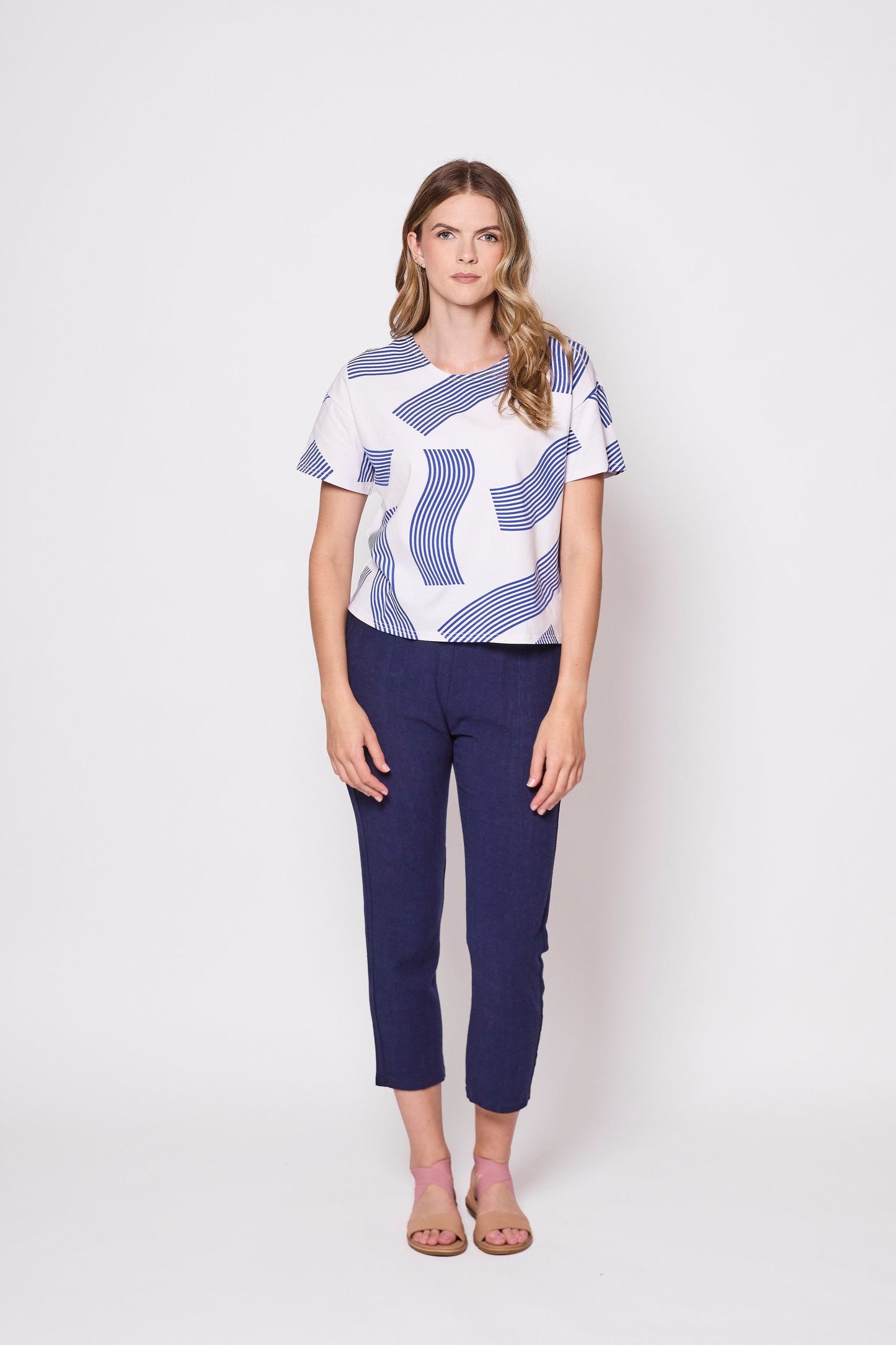 front view of a woman wearing the Waverly T-Shirt by Tangente in Blue, a slightly boxy tee with a wavy striped print on a white background. She is wearing it with blue pants and standing in front of a white background. 