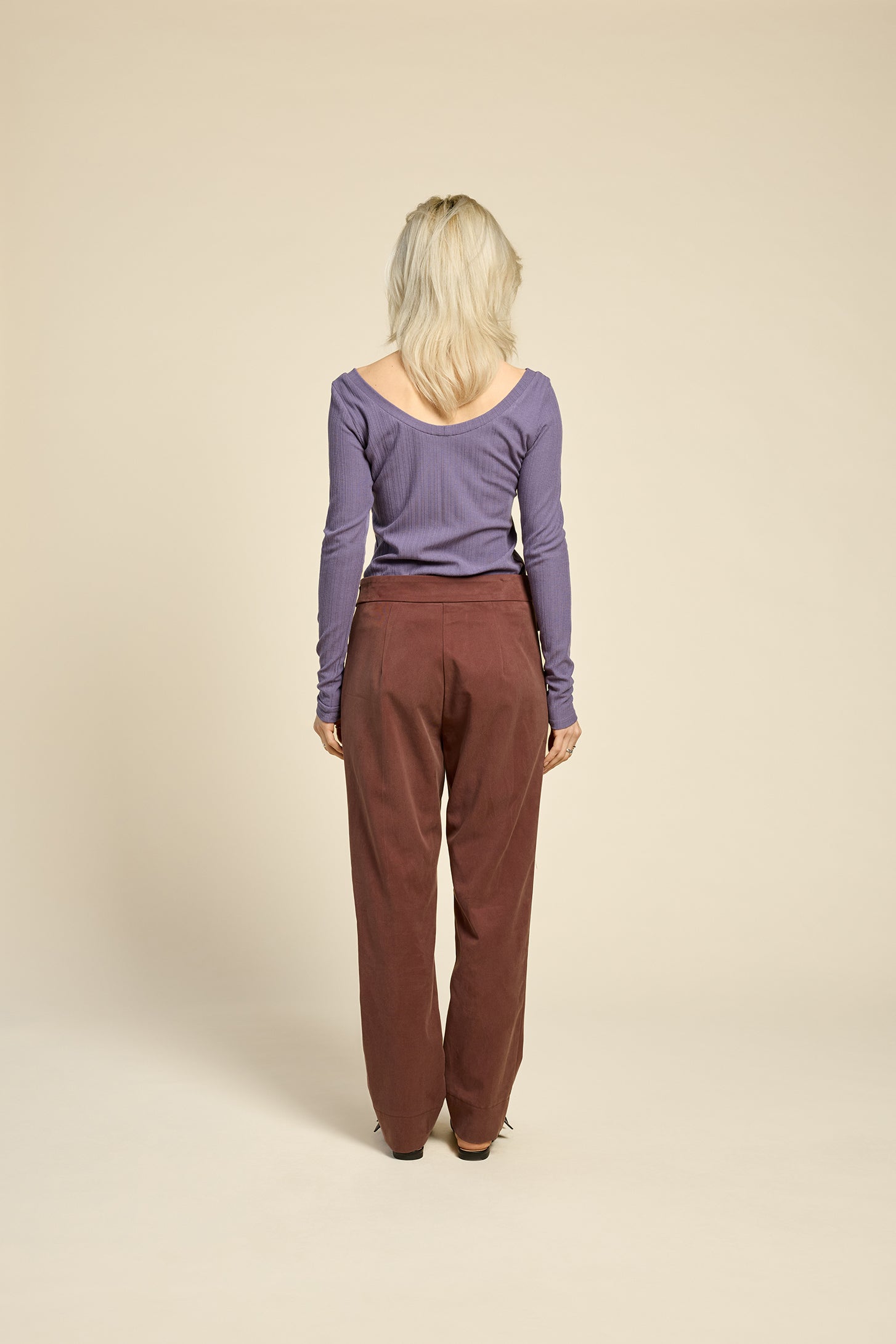 Back view of a woman wearing the Fool Pants by Cokluch in Cappucino, featuring an attached belt with D-rings, a flap and button detail at the waist, and tapered ankle-length legs. She is wearing them with a purple top and standing in front of a beige background