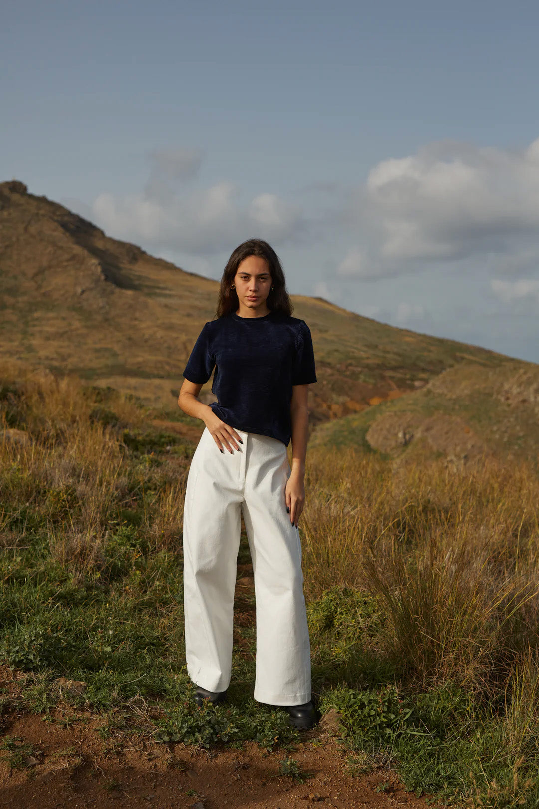 A woman wearing the Tofino Top by MAS in Indigo, a slightly cropped tee in velvet rib. She is wearing it with white pants and is standing on a hill. 