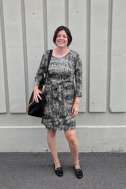 A woman wearing the Gwen Dress by Tangente in Green print, standing outside in front of a white wall
