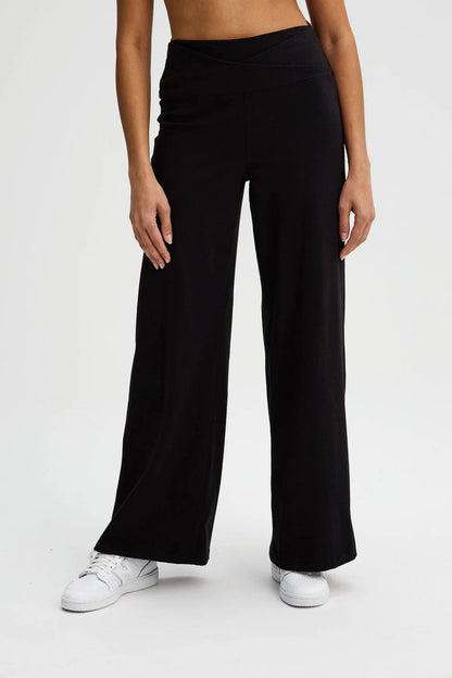 Waist down view of a woman wearing the Villeray Palazzo Pants by Message Factory in Black, wide legged organic cotton pants with a pull-on elastic waistband with a cross-over detail. She is standing in front of a white background. 