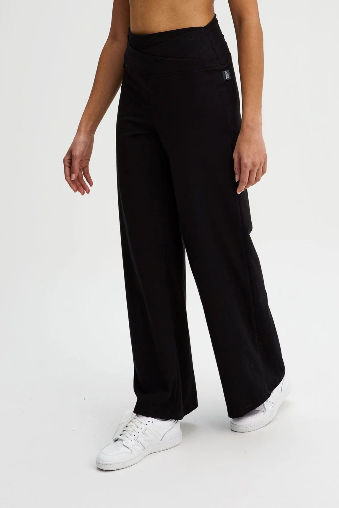 Waist down view of a woman wearing the Villeray Palazzo Pants by Message Factory in Black, wide legged organic cotton pants with a pull-on elastic waistband with a cross-over detail. She is standing in front of a white background. 