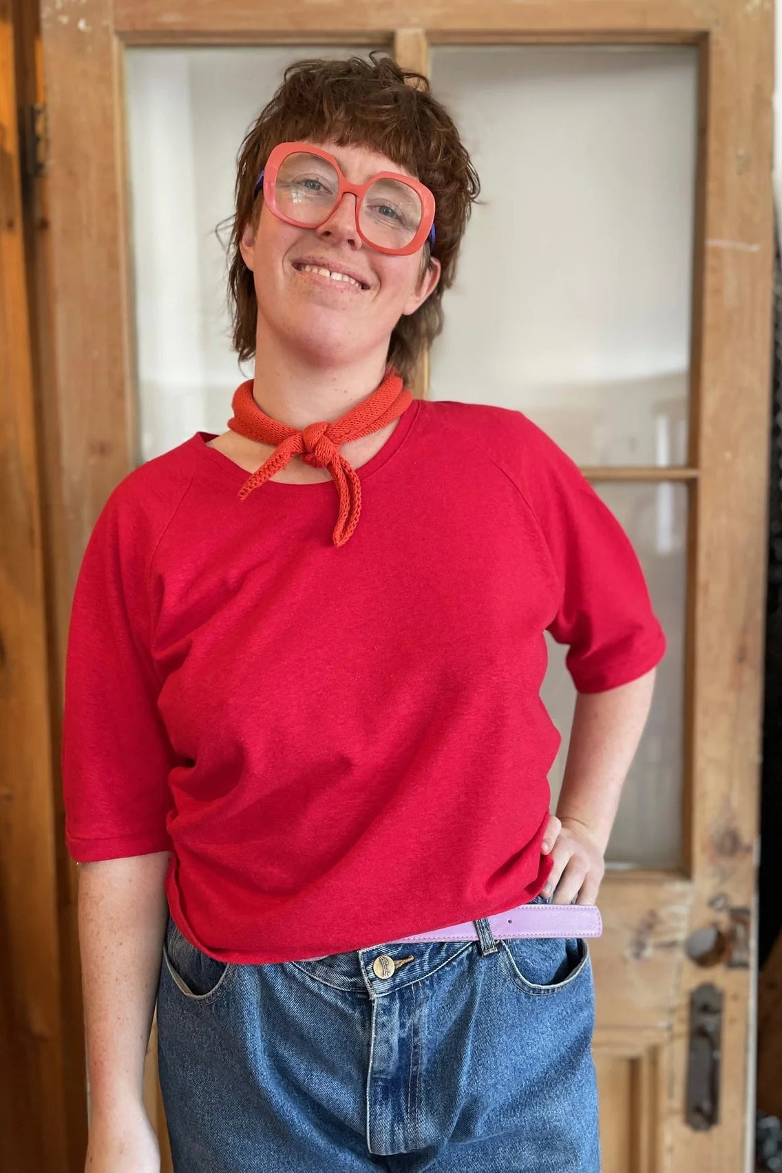 A woman wearing the Vernus Top by Kazak in Tomato with elbow-length raglan sleeves with a pleat detail at the shoulders, a round neck, and a fit that tapers at the waist. She is wearing it with light jeans and an orange scarf and standing indoors by a glass-panelled door. 