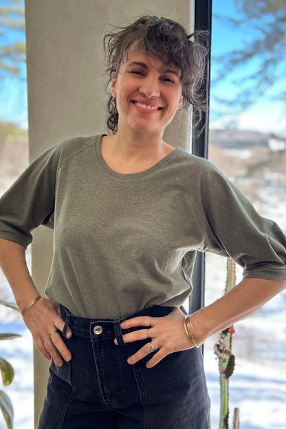 A woman wearing the Vernus Top by Kazak in Sage with elbow-length raglan sleeves with a pleat detail at the shoulders, a round neck, and a fit that tapers at the waist. She is wearing it with dark jeans standing indoors by a window.