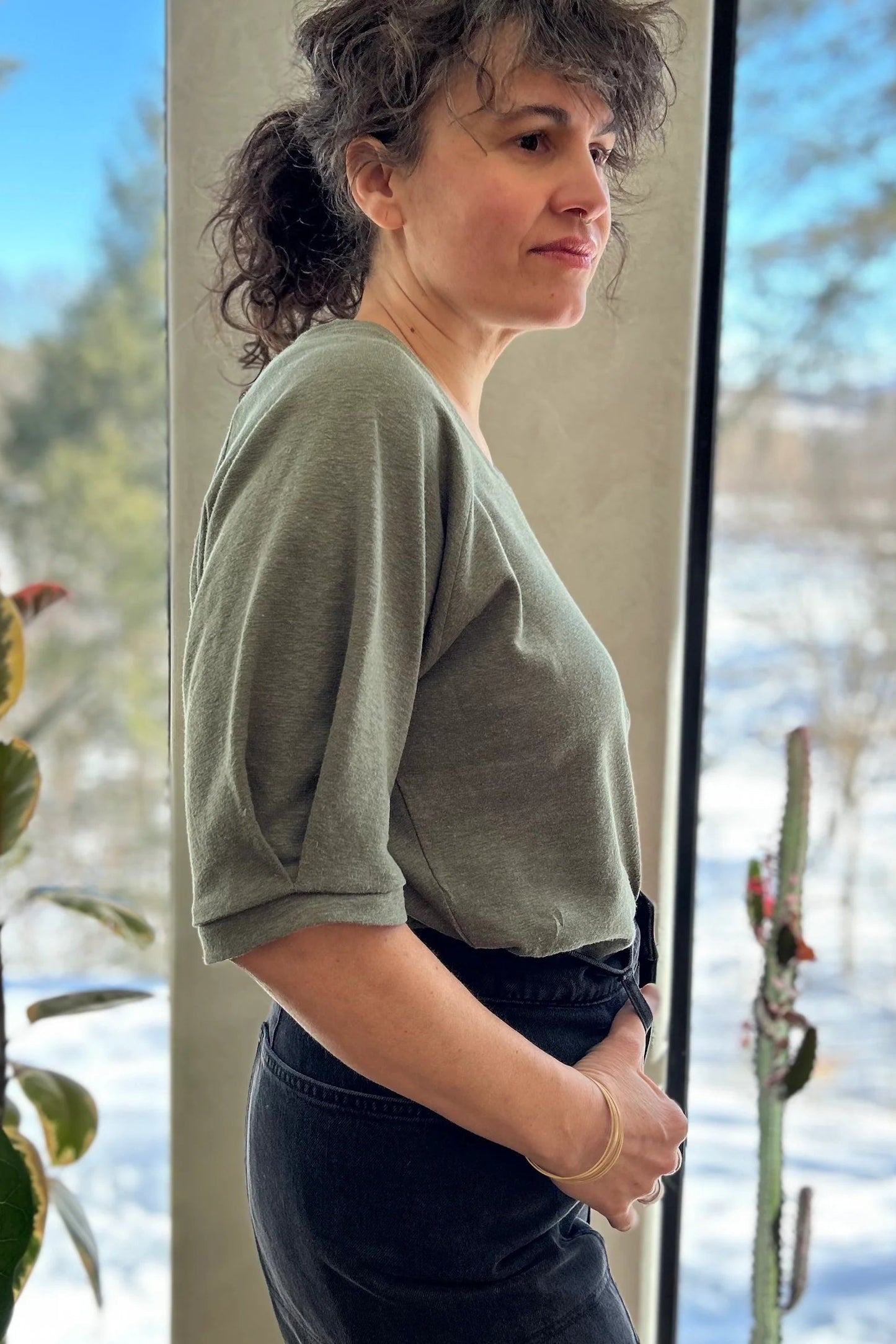 Side view of a woman wearing the Vernus Top by Kazak in Sage with elbow-length raglan sleeves with a pleat detail at the shoulders, a round neck, and a fit that tapers at the waist. She is wearing it with dark jeans standing indoors by a window.
