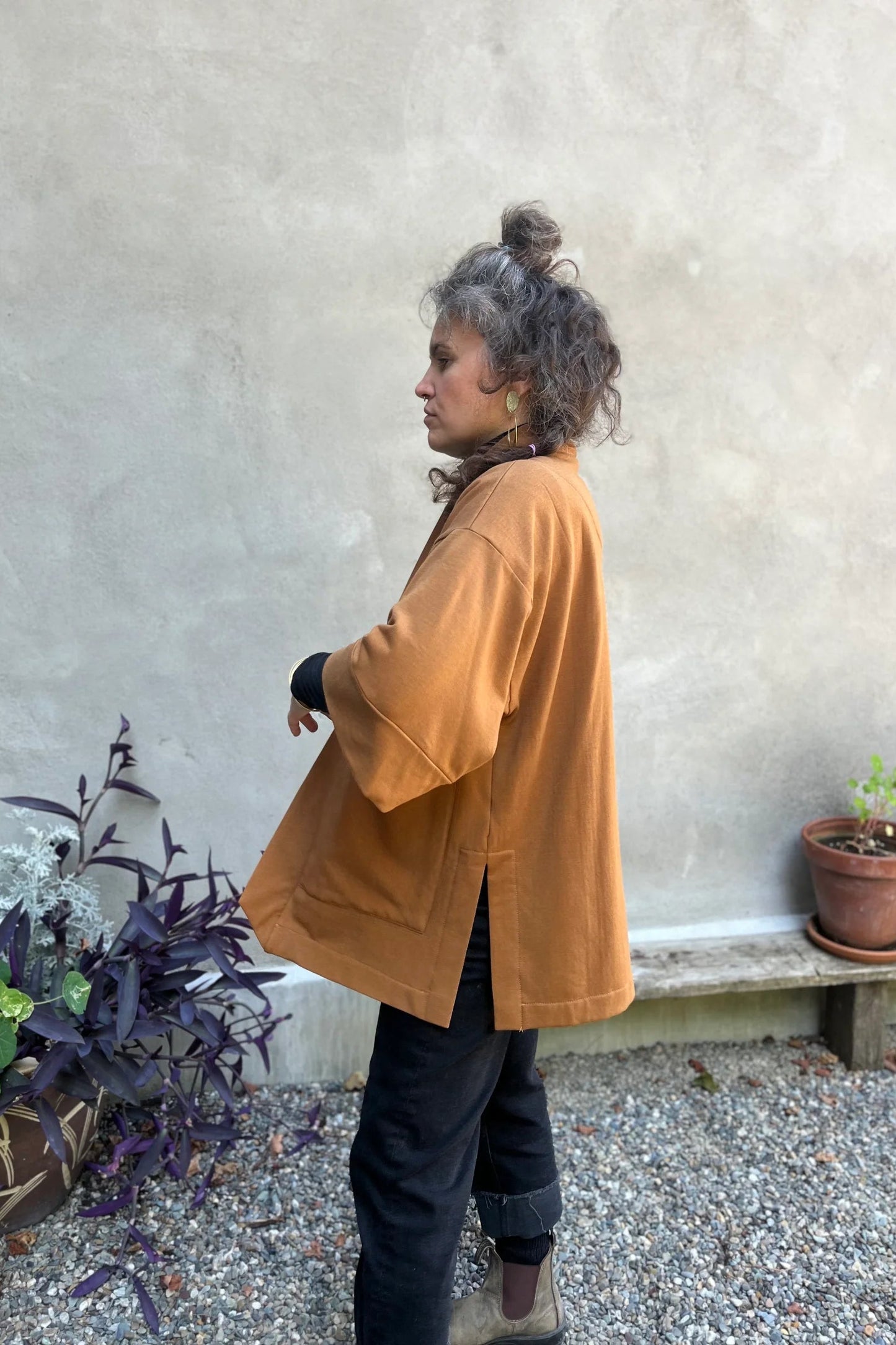 Side view of a woman wearing the Cydonia Cardigan by Kazak in Teak, a hip-length open cardigan with 3/4 kimono sleeves and large patch pockets. She is wearing it with black pants and a black top and standing outside.