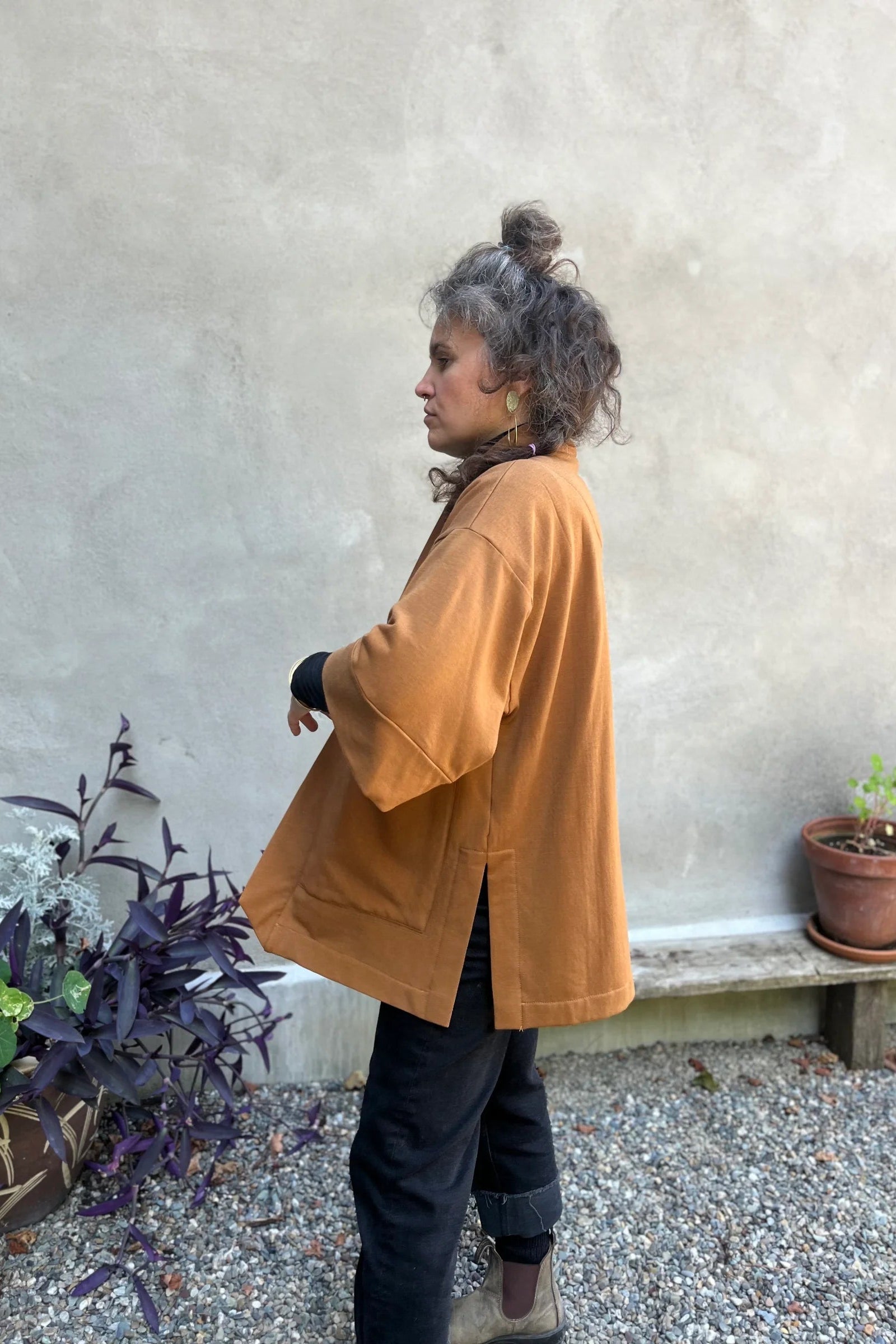 Side view of a woman wearing the Cydonia Cardigan by Kazak in Teak, a hip-length open cardigan with 3/4 kimono sleeves and large patch pockets. She is wearing it with black pants and a black top and standing outside.