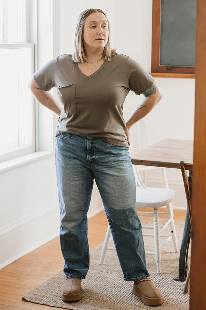 A woman wearing the Wave Tee by Blondie Apparel in Clay with a pair of jeans, standing in front of a table and chairs 