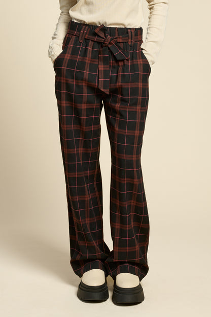 Waist down view of a woman wearing the West Coast Pants by Cokluch in Burgundy Plaid, featuring flared legs, an attached belt with a D-ring closure, and pointed belt loops. She is wearing them with a beige top and standing in front of a beige background.