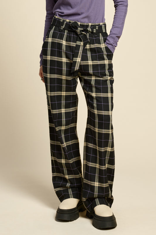 Waist down view of a woman wearing the West Coast Pants by Cokluch in Black Plaid, featuring flared legs, an attached belt with a D-ring closure, and pointed belt loops. She is wearing them with a purple top and standing in front of a beige background.