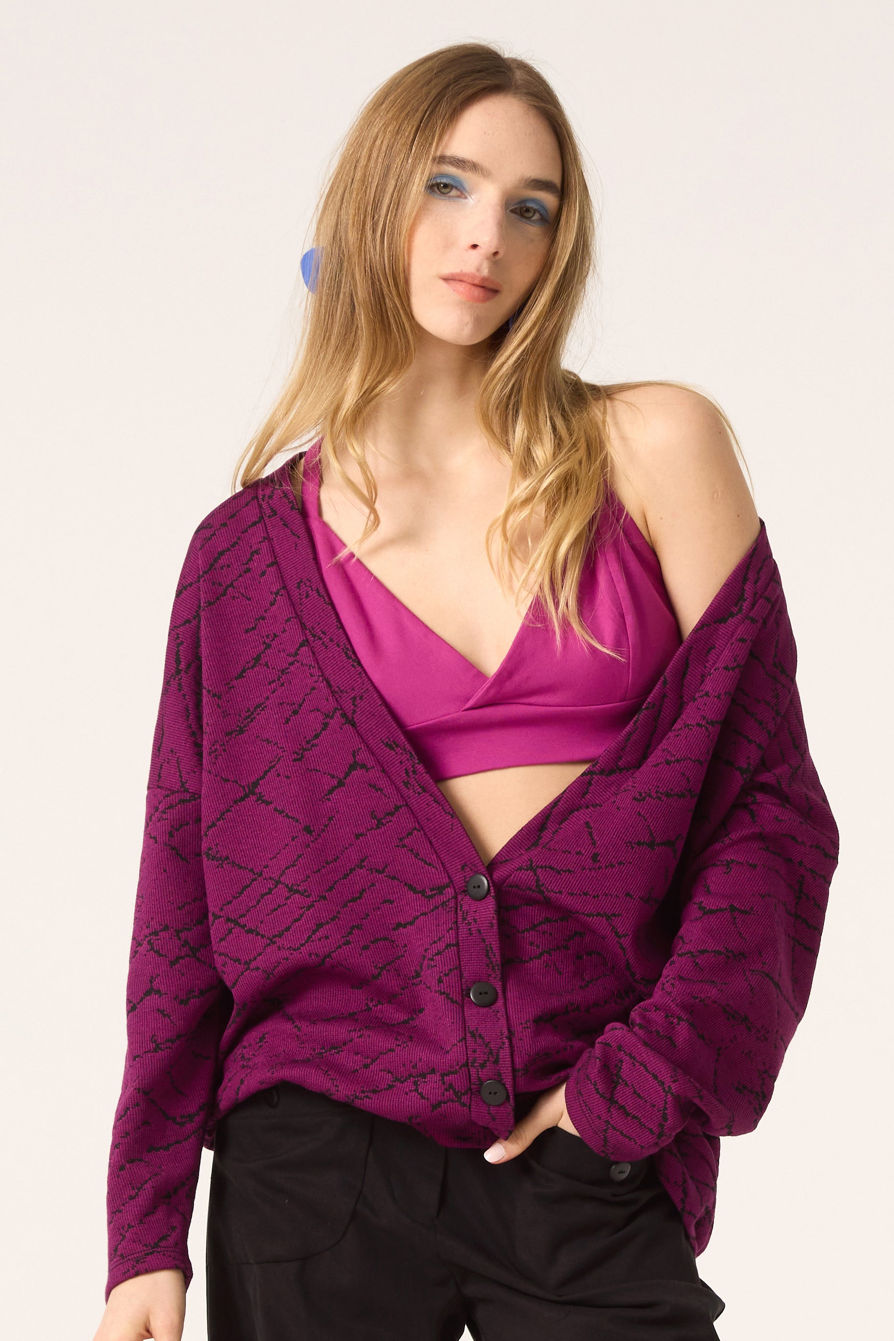 Semi close-up of a woman wearing the Woodland Cardigan by Cokluch in Magenta, with a pink bralette and dark pants, standing in front of a white background. 