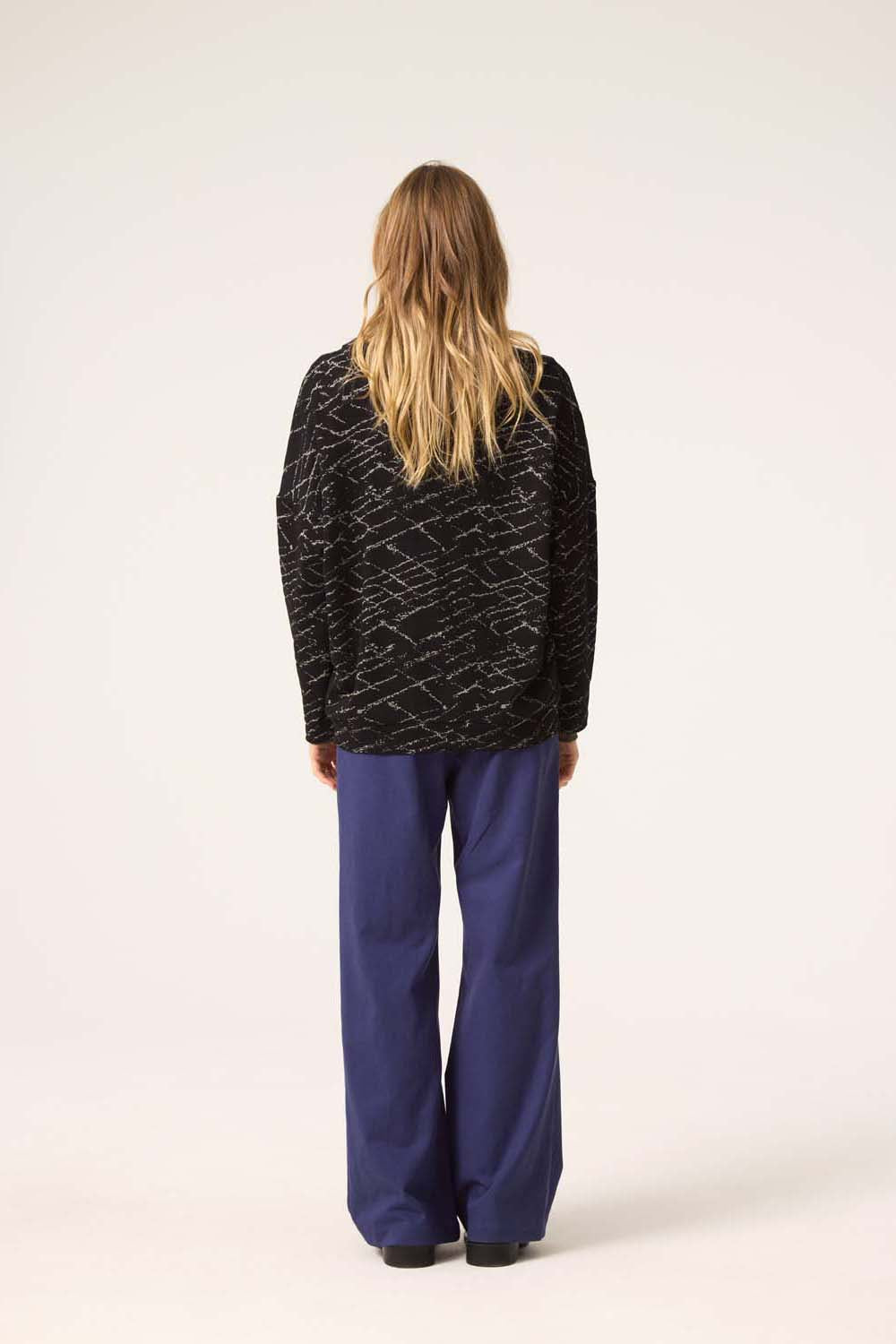 Back view of a woman wearing the Woodland Cardigan by Cokluch in Black, with blue pants, standing in front of a white background. 