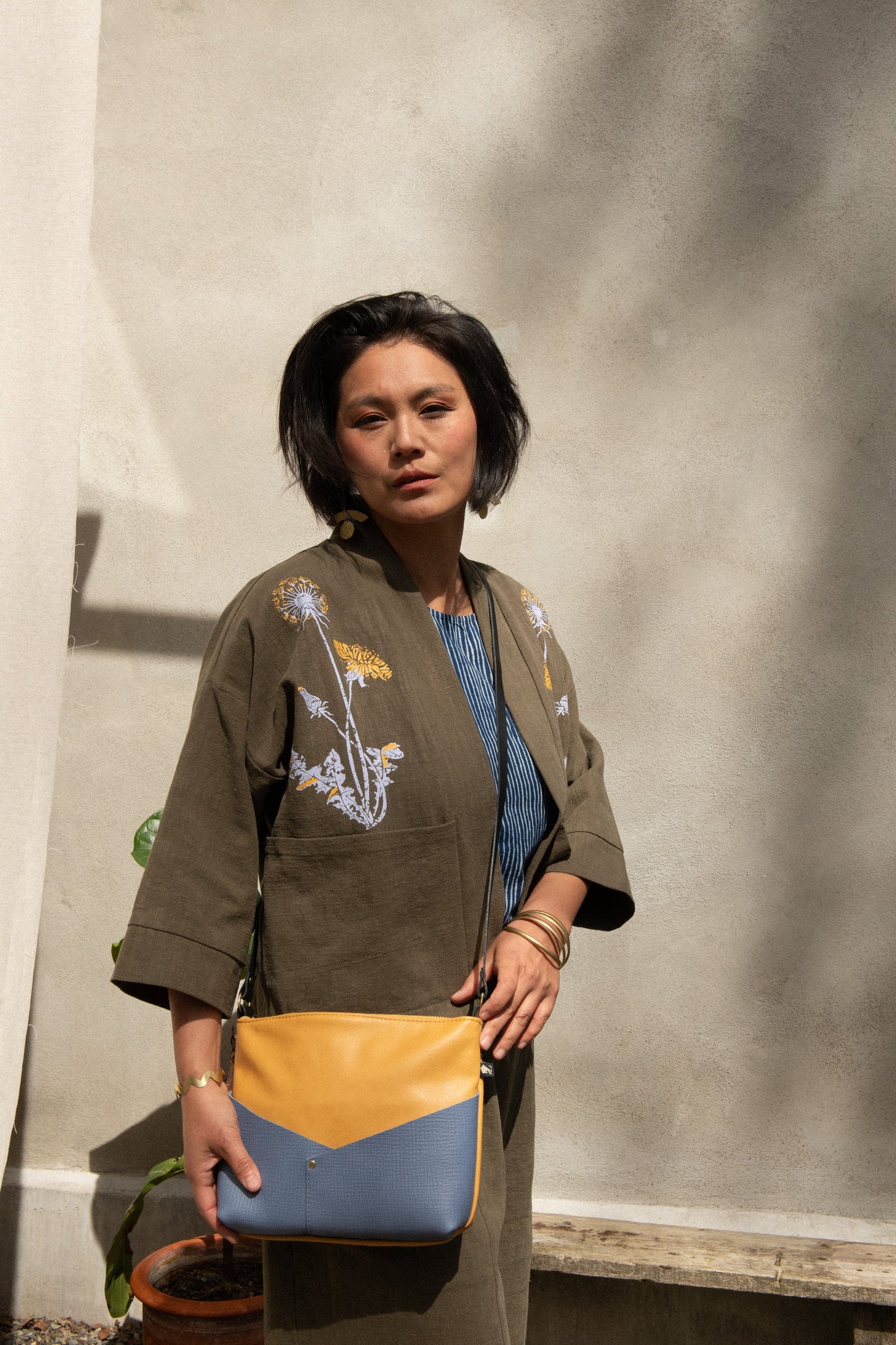 Side view of a woman wearing the Magnolia Screen-printed Jacket from Kazak in Olive with Dandelions, a short kimono style jacket with hand-screen printed dandelions, open front, 3/4-length sleeves, and big patch pockets. She is wearing it with matching pants and a blue and yellow purse and standing outside by a grey wall.